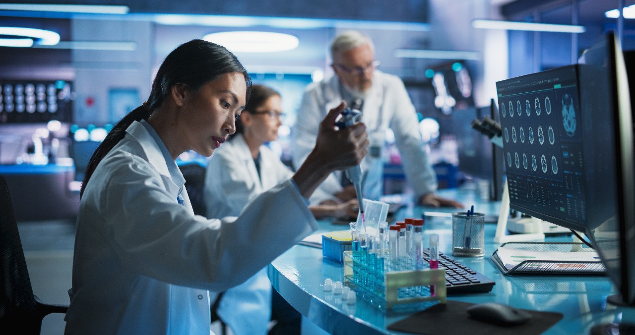 Pharmaceutical Research Laboratory: Female Asian Doctor Working With Test Tubes To Study Innovative Mental Disorder Medicine. Focus Shifting To Caucasian Colleague Looking At CT Brain Scan On Computer
