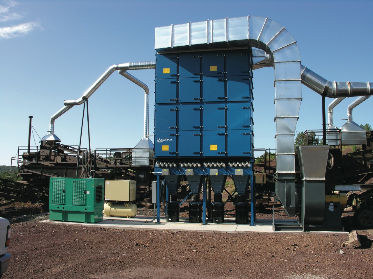 Donaldson Torit® Dalamatic®バグハウス中央集塵機は、コンベヤー移送点の粉塵発生源にダクトで接続します。この形式のバグハウスは、フィルターを圧縮空気でクリーニングするため、集塵機の横に空気コンプレッサー（緑色の装置）と圧縮空気ドライヤー（黄褐色の装置）を設置することが必須でした。